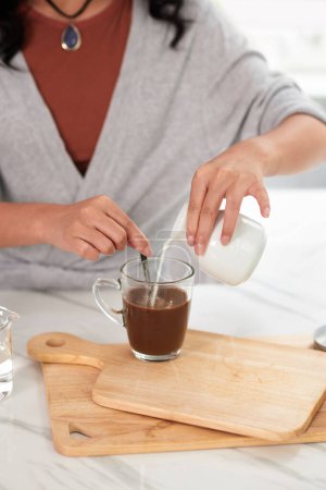 Téléchargez les photos : Gros plan de la jeune femme qui ajoute du lait dans sa tasse avec du café alors qu'elle est assise à table - en image libre de droit