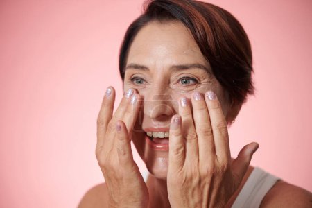Foto de Emocionada mujer madura aplicando crema hidratante en su cara después de la ducha - Imagen libre de derechos