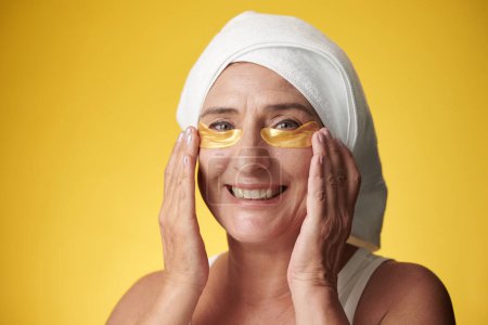Téléchargez les photos : Portrait de femme mûre joyeuse appliquant des patchs hydrogel doré pour réduire les poches et les cernes - en image libre de droit