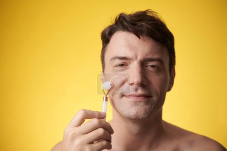 Photo for Middle-aged man enjoying massaging face with roller, isolated on yellow - Royalty Free Image