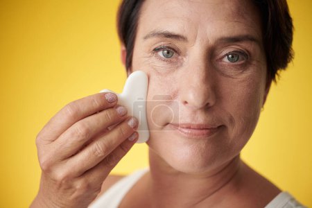 Foto de Mujer de mediana edad masajeando la cara con gua sha para reducir la hinchazón - Imagen libre de derechos