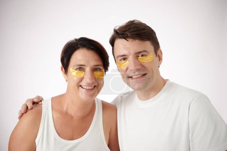 Téléchargez les photos : Couple heureux avec hydrogel sous les patchs des yeux étreignant et souriant à la caméra - en image libre de droit
