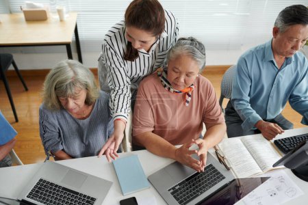 Téléchargez les photos : Jeune femme aidant les femmes âgées confuses à lancer le programme sur les ordinateurs portables - en image libre de droit
