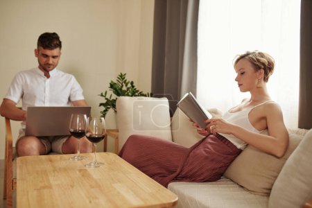 Photo for Young couple reading a book on sofa with her boyfriend using laptop in background while they resting in living room - Royalty Free Image