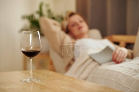Foto de Primer plano de la copa de vino tinto de pie en la mesa con la mujer descansando en el sofá en la habitación - Imagen libre de derechos