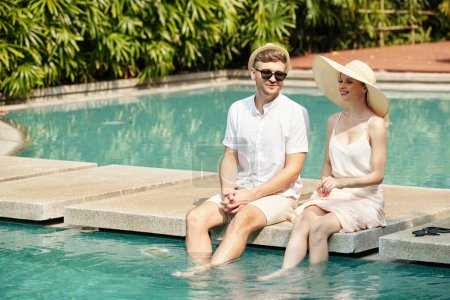 Téléchargez les photos : Jeune couple assis près de la piscine extérieure et parlant, ils passent du temps à la station - en image libre de droit