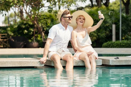 Photo for Young happy couple taking selfie together on mobile phone while sitting at pool at resort - Royalty Free Image