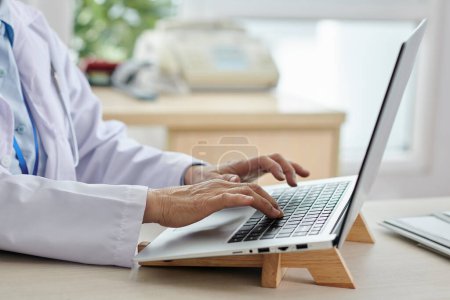 Téléchargez les photos : Gros plan d'une femme médecin assise sur son lieu de travail au bureau et tapant sur un ordinateur portable faisant du travail en ligne - en image libre de droit