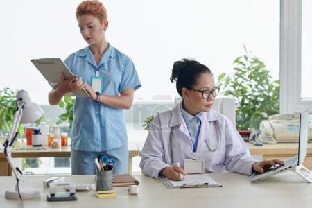 Foto de Médico asiático sentado en la mesa y trabajando en el ordenador portátil con la enfermera examinando la tarjeta médica del paciente en segundo plano - Imagen libre de derechos