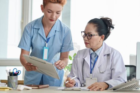 Téléchargez les photos : Jeune infirmière discutant de la carte patient avec le médecin pendant qu'ils travaillent au cabinet médical - en image libre de droit