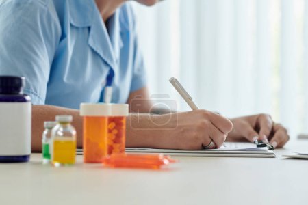 Foto de Primer plano de la enfermera joven recetando medicamentos para el paciente mientras está sentada en la mesa con frascos de pastillas - Imagen libre de derechos