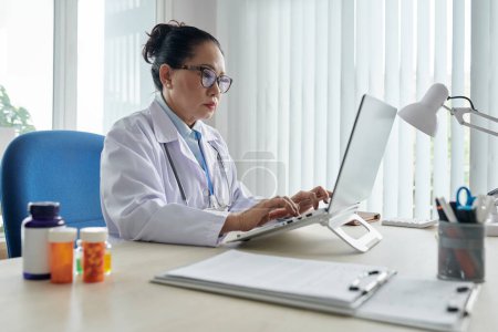 Téléchargez les photos : Asiatique femme médecin dans les lunettes de dactylographie sur ordinateur portable faire du travail en ligne à la table au bureau - en image libre de droit