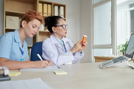 Téléchargez les photos : Médecin asiatique utilisant un ordinateur portable pour présenter un nouveau médicament lors d'une réunion en ligne pendant que l'infirmière prend des notes sur la carte à la table - en image libre de droit