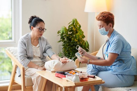 Foto de Enfermera joven examinando la presión arterial del paciente con equipo médico durante la visita domiciliaria - Imagen libre de derechos