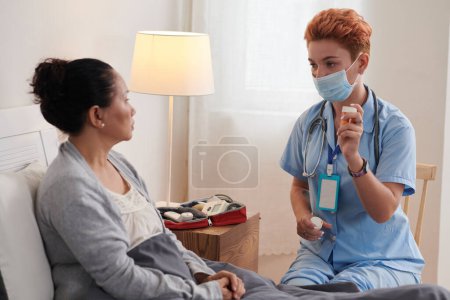 Photo for Young nurse in mask prescribing medicine for treatment to sick patient in bed during home visit - Royalty Free Image
