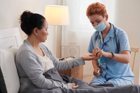 Foto de Enfermera joven en uniforme dando píldora a una anciana enferma mientras está sentada en la cama - Imagen libre de derechos