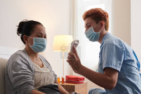 Téléchargez les photos : Jeune infirmière utilisant un équipement médical pour mesurer la température de la femme âgée - en image libre de droit