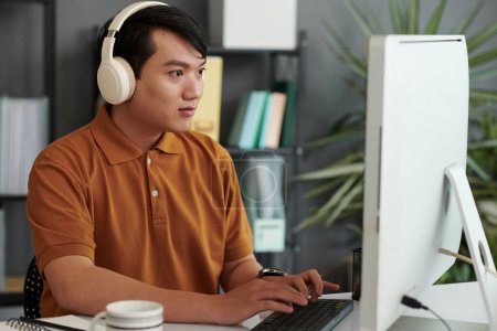 Photo for Young Vietnamese programmer listening to music in headphones when working on computer - Royalty Free Image