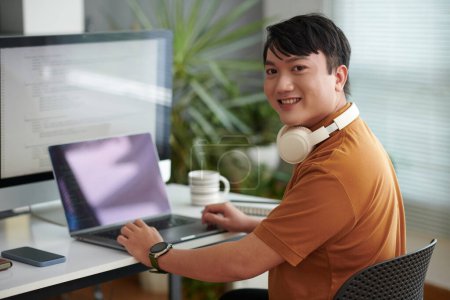 Téléchargez les photos : Portrait de joyeux développeur de logiciels asiatiques travaillant sur ordinateur à son bureau et se tournant vers la caméra - en image libre de droit