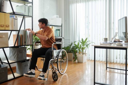 Foto de Joven con discapacidad desplazándose por casa oficina en silla de ruedas en busca de documentos - Imagen libre de derechos