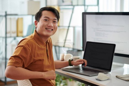 Photo for Portrait of cheerful senior developer sitting at desk with computer and laptop with programming code on screens - Royalty Free Image