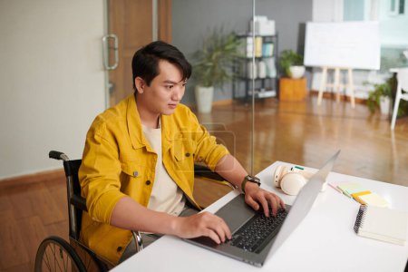 Téléchargez les photos : Développeur de logiciels vietnamien en fauteuil roulant travaillant sur ordinateur portable au bureau - en image libre de droit