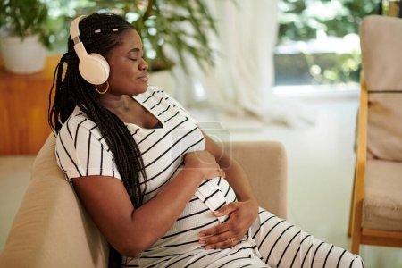 Foto de Calma mujer negra cerrando los ojos y escuchando música de embarazo para la madre y el bebé nonato - Imagen libre de derechos