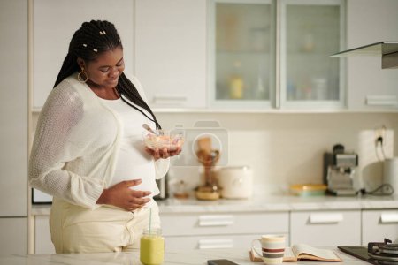 Photo for Smiling pregnant woman eating healthy breakfast and touching her belly - Royalty Free Image