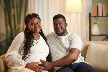 Photo for Portrait of smiling husband and his pregnant wife sitting on couch at home - Royalty Free Image