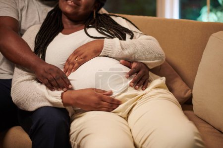 Foto de Embarazada mujer negra acariciando con marido en sofá en casa - Imagen libre de derechos