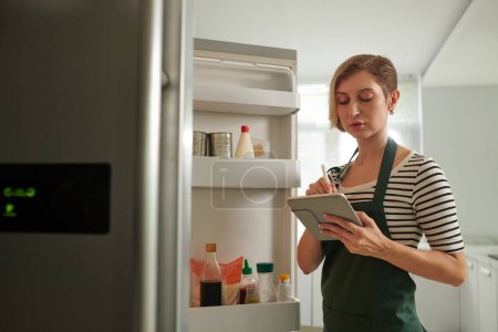 Téléchargez les photos : Femme debout devant le réfrigérateur ouvert et remplissant la liste d'achats sur tablette - en image libre de droit
