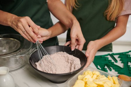 Photo for Mother showing teenage daughter how to use whisk when mixing cake ingredients together - Royalty Free Image