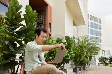 Photo for Young businessman sitting in front of office building, opening laptop to answer e-mails - Royalty Free Image