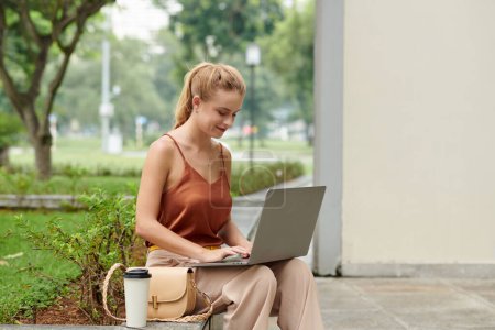 Photo for Smiling young woman sitting on bench in park and working on laptop - Royalty Free Image