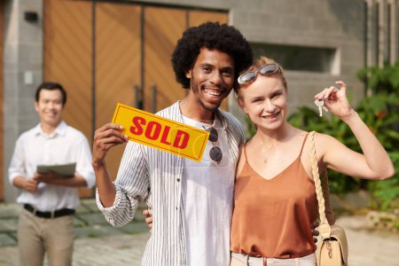 Foto de Retrato de feliz pareja diversa excitada mostrando letrero vendido y llaves de casa nueva - Imagen libre de derechos