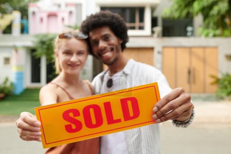 Foto de Vendido letrero en manos de feliz pareja diversa que compró nueva casa - Imagen libre de derechos