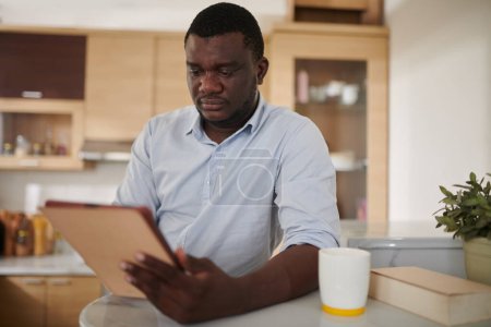 Téléchargez les photos : Portrait d'un homme debout dans la cuisine, buvant du café et lisant un livre électronique sur tablette - en image libre de droit