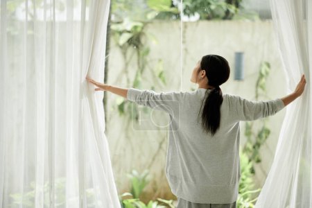 Foto de Mujer joven abriendo cortinas de gran ventana, vista desde atrás - Imagen libre de derechos