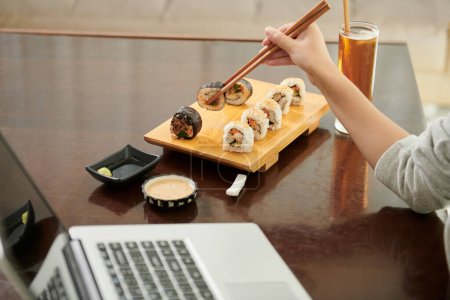 Photo for Cropped image of female freelancer eating sushi in cafe when working on laptop - Royalty Free Image