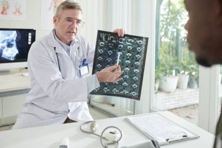 Photo for Serious doctor explaining patient why he has to care about spine health and pointing at discs MRI image - Royalty Free Image