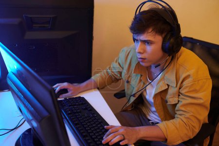 Téléchargez les photos : Jeune homme sérieux portant un casque lors de la lecture de jeux vidéo sur ordinateur à la maison - en image libre de droit