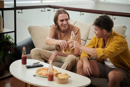 Téléchargez les photos : Des copains souriants boivent des bières et mangent des pizzas à la maison - en image libre de droit