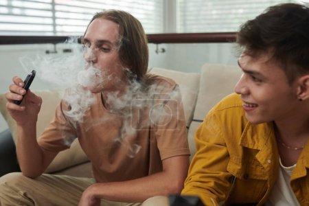 Téléchargez les photos : Jeune homme fumant vape lors de passer du temps avec le meilleur ami - en image libre de droit