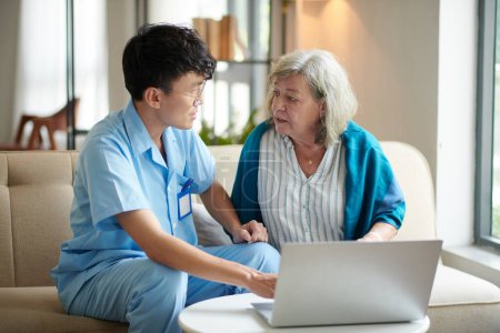 Foto de Mujer mayor que pide a la trabajadora médica que le explique cómo usar el servicio de telemedicina en la computadora - Imagen libre de derechos