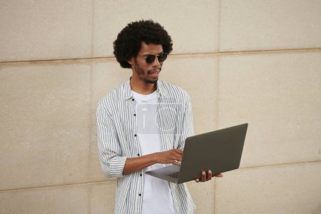 Téléchargez les photos : Portrait de jeune homme noir fronçant les sourcils dans des lunettes envoyer des e-mails aux clients - en image libre de droit