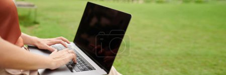 Téléchargez les photos : Mains de pigiste assis sur la prairie dans le parc et travaillant sur ordinateur portable - en image libre de droit