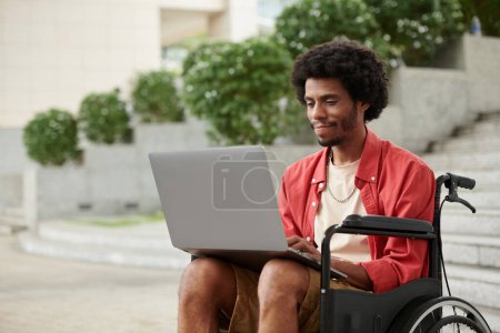 Foto de Joven hombre negro con discapacidad que trabaja como freelancer, él está respondiendo correos electrónicos en el ordenador portátil - Imagen libre de derechos