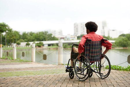 Téléchargez les photos : Jeune homme en fauteuil roulant passer du temps sur la rive de la rivière profiter de la vue sur la ville - en image libre de droit