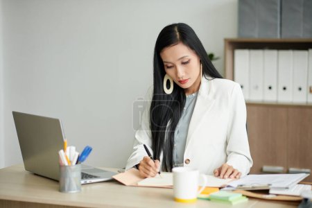 Foto de Retrato de una mujer empresaria elegante y seria que trabaja con documentos en el escritorio de su oficina y toma notas en el planificador - Imagen libre de derechos