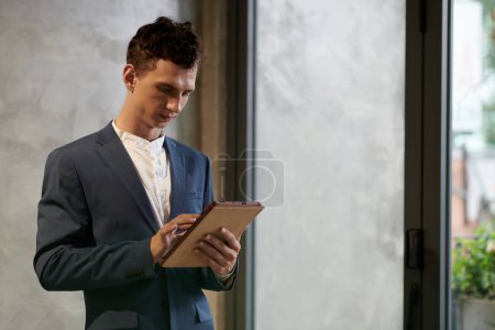 Téléchargez les photos : Sérieux jeune entrepreneur debout dans la salle de bureau et de lire l'article sur tablette - en image libre de droit